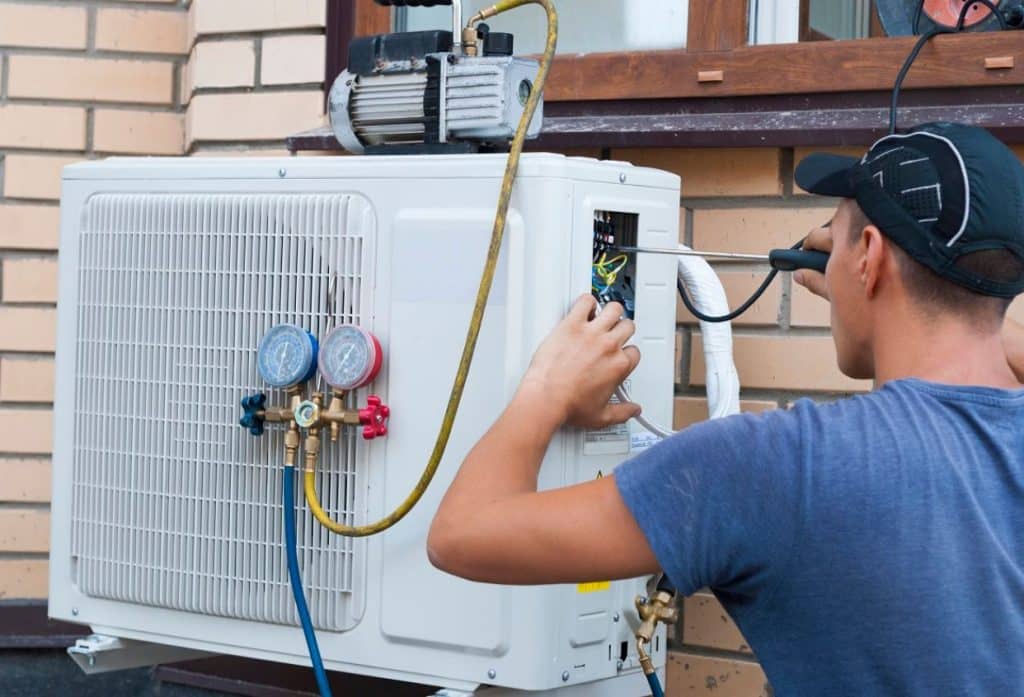 A technician services an outdoor air-conditioning unit, checking its electrical components while connected to refrigerant gauges and hoses.