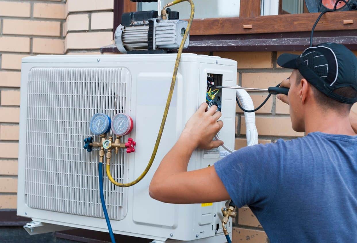 A technician services an outdoor air-conditioning unit, checking its electrical components while connected to refrigerant gauges and hoses.
